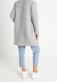 Cappotto di lana grigio con taglio dritto, maniche lunghe e finitura texturizzata, abbinato a pantaloni cropped azzurri e sneakers bianche.