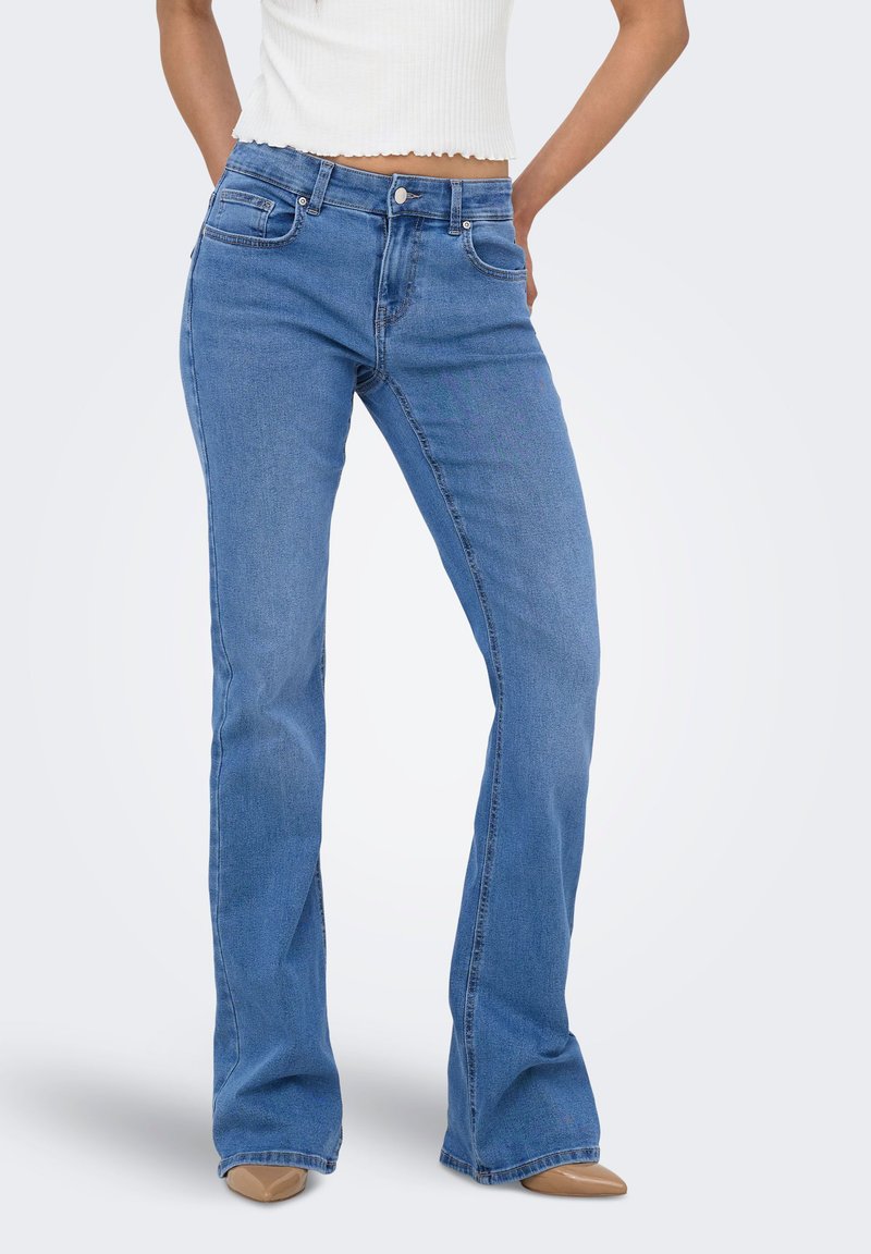 Γυναίκα με ανοιχτό μπλε τζιν bootcut, λευκό κοντό τοπ και καφέ μυτερές γόβες, στέκεται με τα χέρια στη μέση.