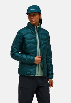 Femme portant une veste matelassée teal par-dessus un sweat à capuche vert clair zippé, un pantalon noir et une casquette teal assortie, regardant sur le côté avec une expression neutre.