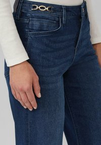 Donkerblauwe spijkerbroek met een gouden gesp op de tailleband, met een gladde textuur en een klassieke pasvorm. Geen decoratieve patronen.