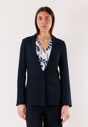 Donna con capelli lisci e castani che indossa un blazer blu navy sopra una camicia bianca con motivi blu, in piedi contro uno sfondo chiaro e uniforme.