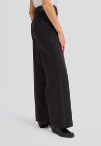 Person wearing high-waist wide-leg black jeans and black square-toe boots, standing sideways against plain background.