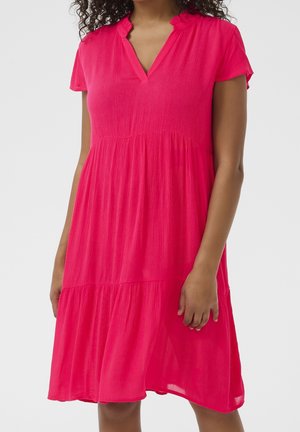 Femme portant une robe rose vif à manches courtes, longueur genou, avec un décolleté en V et un col à volants, debout devant un fond uni.