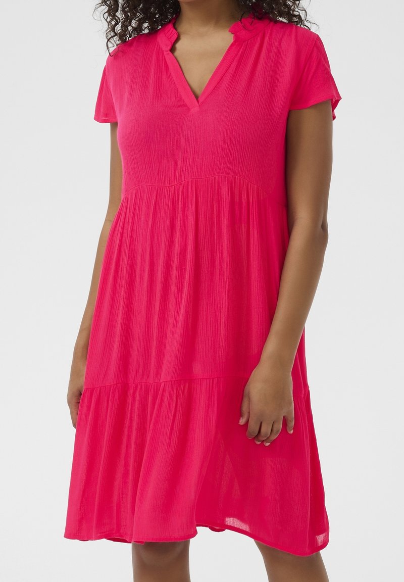 Femme portant une robe rose vif à manches courtes, longueur genou, avec un décolleté en V et un col à volants, debout devant un fond uni.