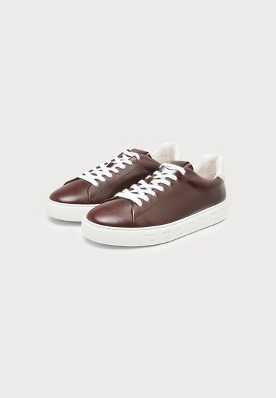 Paire de baskets en cuir marron avec des lacets blancs et des semelles blanches épaisses comportant des motifs embossés sur un fond gris clair.
