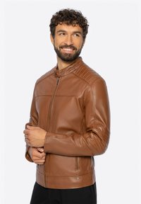 Usměvavý muž s tmavými kudrnatými vlasy a vousy, oblečený v hnědé kožené bundě na zip a černých kalhotách, hledící doprava.