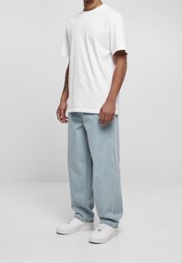 Uomo che indossa una maglietta bianca semplice, jeans larghi azzurri chiari e sneakers bianche, in piedi contro uno sfondo grigio neutro.