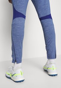 Blauwe sportbroek van textuurstof. Heeft paarse accenten en ritsbuiken. Draagt met witte sneakers die neon details accentueren.