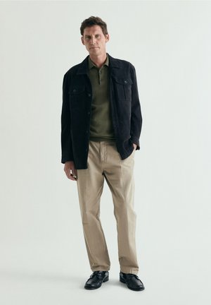 Man wearing black jacket over olive green polo shirt, beige trousers, and black shoes, standing against plain light background.