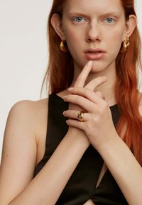 Anillo de oro con diseño en espiral que se lleva en los dedos; el modelo muestra una piel texturizada, usando pendientes de aro de oro con un atuendo negro.
