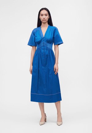 Femme aux longs cheveux foncés portant une robe midi bleue à manches courtes et des talons beiges, debout devant un fond clair uni.