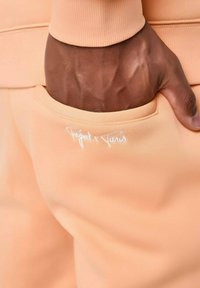 Jogging peach con una textura suave, que presenta un bolsillo lateral y un logo bordado en blanco. Confeccionado con una tela lisa y elástica.