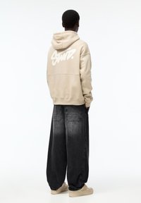 Beige Kapuzenpullover mit großem weißen Logo auf dem Rücken, kombiniert mit lockeren, dunklen Denim-Hosen. Das Model steht mit dem Rücken zum Betrachter und zeigt einen lässigen Streetwear-Stil.