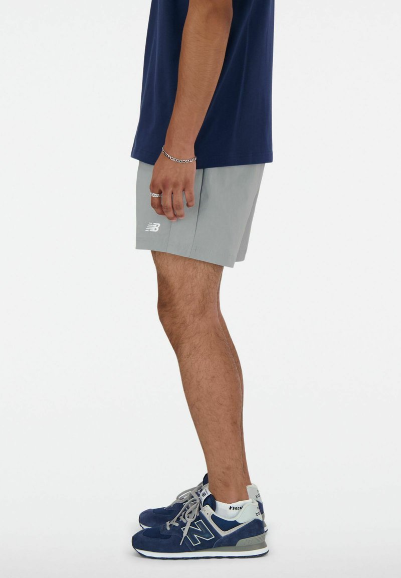 Shorts deportivos gris claro con cintura elástica y logotipo bordado. Combinados con zapatillas de deporte azul marino conacentos grises y marcas blancas.
