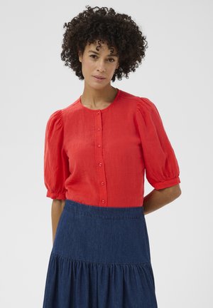 Femeie cu păr creț, purtând o bluză roșie cu nasturi, mâneci bufante și o fustă albastru închis, cu talie înaltă, pe un fundal simplu.