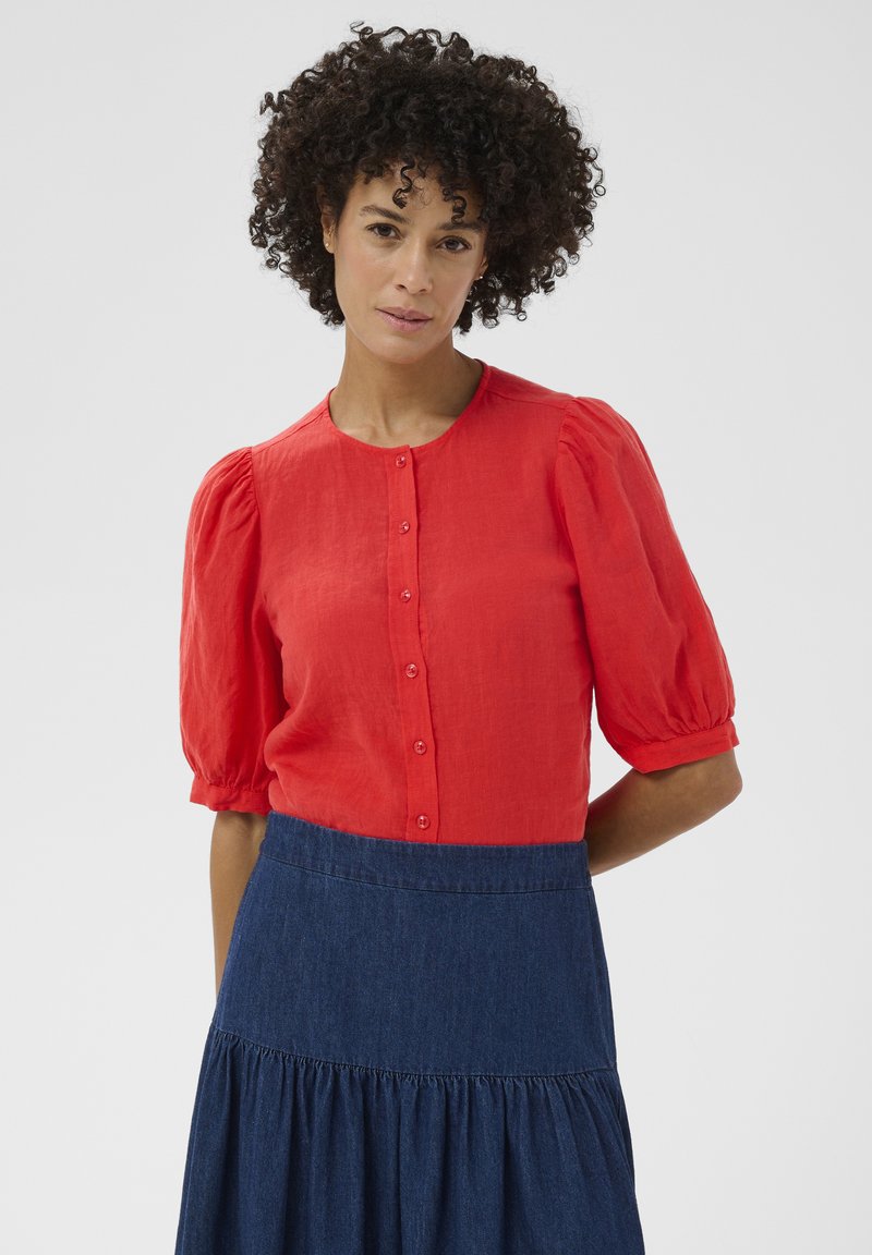 Femme aux cheveux bouclés portant un chemisier rouge à boutons avec des manches bouffantes et une jupe bleu foncé taille haute sur un fond uni.