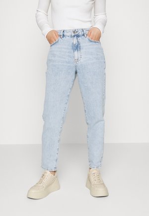 Persoon draagt lichtblauwe hooggetailleerde jeans, wit geribd longsleeve topje en beige platformsneakers, staand met handen in de zakken.