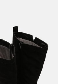 Bottes noires montant jusqu'aux genoux avec extérieur en daim souple, fermeture éclair et doublure en tissu. Elles comportent un logo en haut et une finition légèrement texturée.