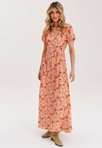 Maxi šaty s kvetinovým vzorom v odtieňoch ružovej a oranžovej, krátke rukávy a pás s nariaseným detailom. Nosené s béžovými čižmami.