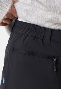 Svarta byxor med elastisk midja, dragkedjeficka och liten blå och gul flaggdetalj. Slät textur och modern design.