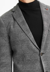 Grå blazer med rutigt mönster, strukturerad design och spetsig slag. Bärs över en svart polotröja, med en knapppdetail.