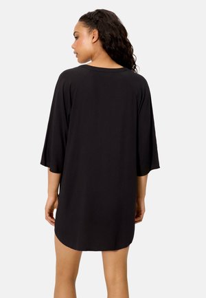 Mujer con cabello largo y rizado, vestida con un vestido negro suelto hasta la rodilla y mangas tres cuartos, vista desde atrás.