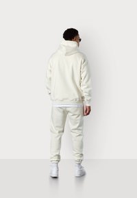 Ensemble sweat-shirt et jogging couleur crème, fabriqué en tissu doux. Dispose d'un col haut, de poignets élastiques et de baskets blanches. Design simpliste.