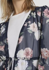 Žena v průhledném kimonu s námořnickými modrými květinovými vzory, které má přes bílý ribovaný top, uvázané v pase na šňůrku ze stejného materiálu.