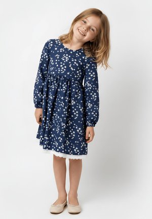 Uśmiechnięta młoda dziewczyna w granatowej sukience z długim rękawem w biały motyw kwiatowy oraz beżowych balerinach, stojąca na białym tle.