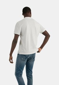 Polo bianco con una texture morbida, maniche corte e bordi a contrasto. Abbinato a jeans blu aderenti, che mostrano la vista posteriore.