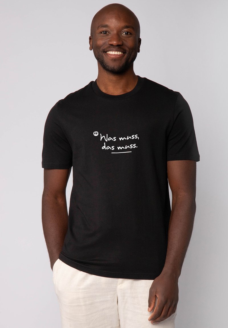 T-shirt en coton noir à manches courtes et col rond. Présente un texte imprimé en blanc "Was muss, das muss" sur le devant.