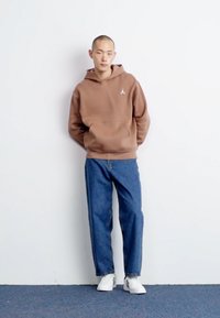 Hoddie marron avec une poche avant et un logo blanc, associé à un jean bleu et des baskets blanches, sur fond blanc uni.