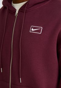 Mørkerød hættejakke med lynlås lavet af blødt stof, med et hvidt broderet Nike-logo på venstre bryst og sølv metal lynlås.