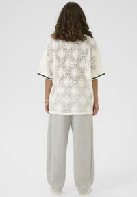 Ženska s kratkimi kodrastimi lasmi, oblečena v belo čipkasto majico s cvetličnimi vzorci in ohlapnimi sivimi hlačami, stoji na enostavni beli podlagi.