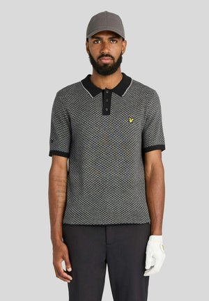 Uomo che indossa una polo grigia con motivo, pantaloni neri, un berretto grigio e un guanto bianco sulla mano destra, in piedi davanti a uno sfondo semplice.