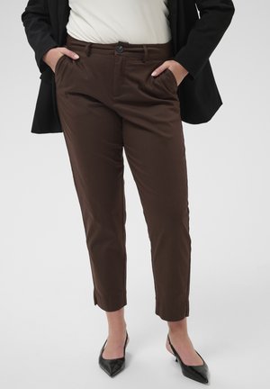Žena nosí hnedé slim-fit skrátené nohavice, čierne slingback topánky so špicatou špičkou, biely top a čierne sako, ruky v vreckách, stojaca.