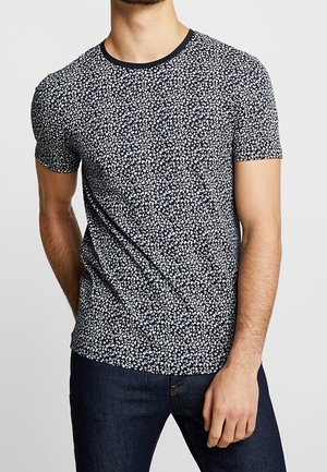Homme portant un t-shirt bleu marine à manches courtes avec un motif floral blanc et un jean bleu foncé, debout avec une main détendue le long du corps.