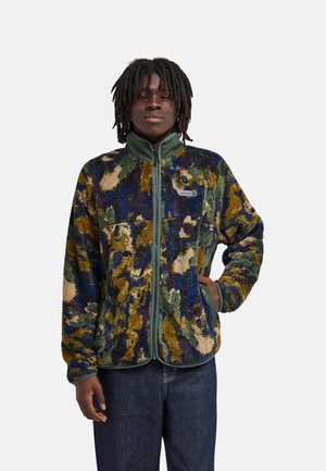 Personne portant une veste polaire multicolore à motif camouflage avec un col et une fermeture éclair verts, debout avec une main dans la poche sur un fond uni.