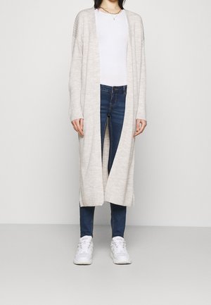 Femme portant un long cardigan beige à ouverture devant sur un t-shirt blanc, un jean skinny bleu, des baskets blanches et des colliers en or, debout devant un mur blanc.