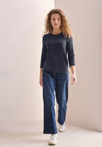 Marineblauer Pullover mit Dreiviertelärmeln, ausgestattet mit einem strukturierten "CREATIVE"-Design, kombiniert mit weit geschnittenen blauen Jeans und weißen Sneakers.