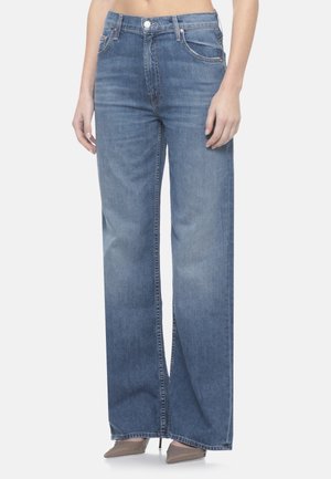 Persona che indossa jeans blu a vita alta e gamba dritta e tacchi beige con punta a punta, in piedi con le mani rilassate lungo i fianchi.