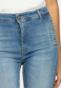 Hoge taille spijkerbroek in lichtblauw met een vijfknoppen sluiting, subtiele vervaging en contraststiksels langs de naden. Soepele textuur.