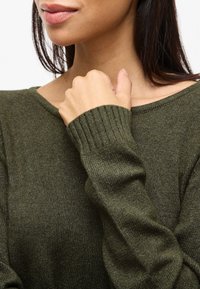 Maglione verde oliva con polsini a costine e scollatura ampia, realizzato in un materiale a maglia testurizzata. Primo piano della scollatura e delle maniche.