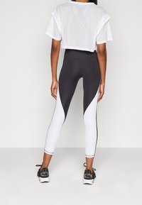 Kobieta ubrana w legginsy w kolorze czarno-białym i białą krótka bluzkę z siateczki, stojąca na tle gładkiego białego tła.