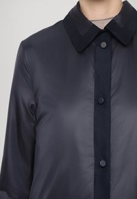 Chemise bleu marine à boutons avec un col classique, partiellement visible sur une personne portant un sous-pull beige clair en dessous.