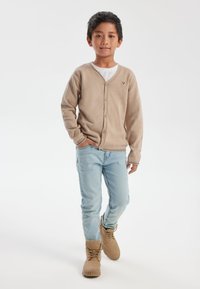 Bež kardigan s V-izrezom i gumbima, nosi se preko bijele majice. Svijetloplave traperice i smeđe čizme upotpunjuju odjevnu kombinaciju.