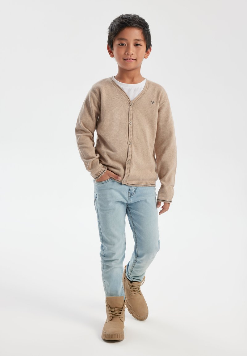 Bež kardigan s V-izrezom i gumbima, nosi se preko bijele majice. Svijetloplave traperice i smeđe čizme upotpunjuju odjevnu kombinaciju.