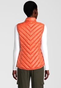Pomarańczowa puchowa kamizelka w jodełkę, wykonana z lekkiego materiału, z wysokim kołnierzem i bez rękawów, zestawiona z białą koszulą z długim rękawem.