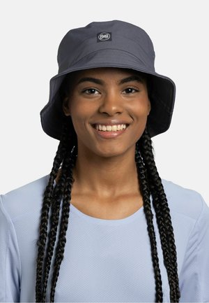 Smiling person with long braids wearing a gray bucket hat with a logo and a light blue textured shirt against a plain background.
