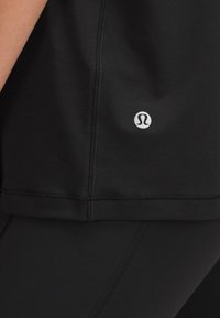 Crna sportska majica s glatkom teksturom, s logotipom na donjoj strani i čistim, minimalističkim dizajnom. Šavovi su vidljivi.